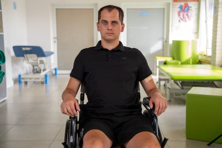 Michał w czarnej koszulce polo siedzi na wózku w sali rehabilitacji i patrzy prosto w obiektyw.