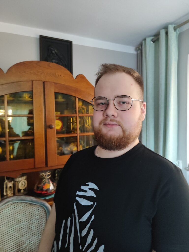 Mikołaj Janczarek w okularach pozuje do zdjęcia. W tle widoczny fragment pokoju z drewnianą komodą.