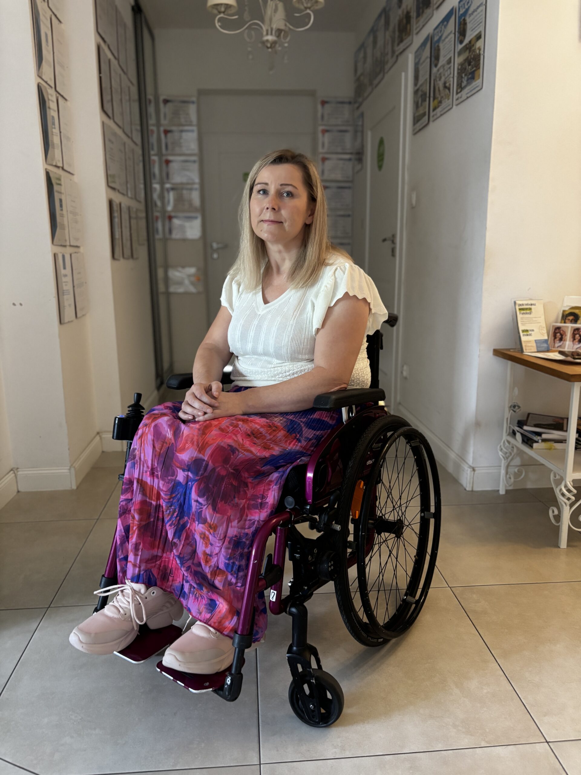 Agnieszka Krukowska siedzi na wózku inwalidzkim pozując na tle korytarza.
