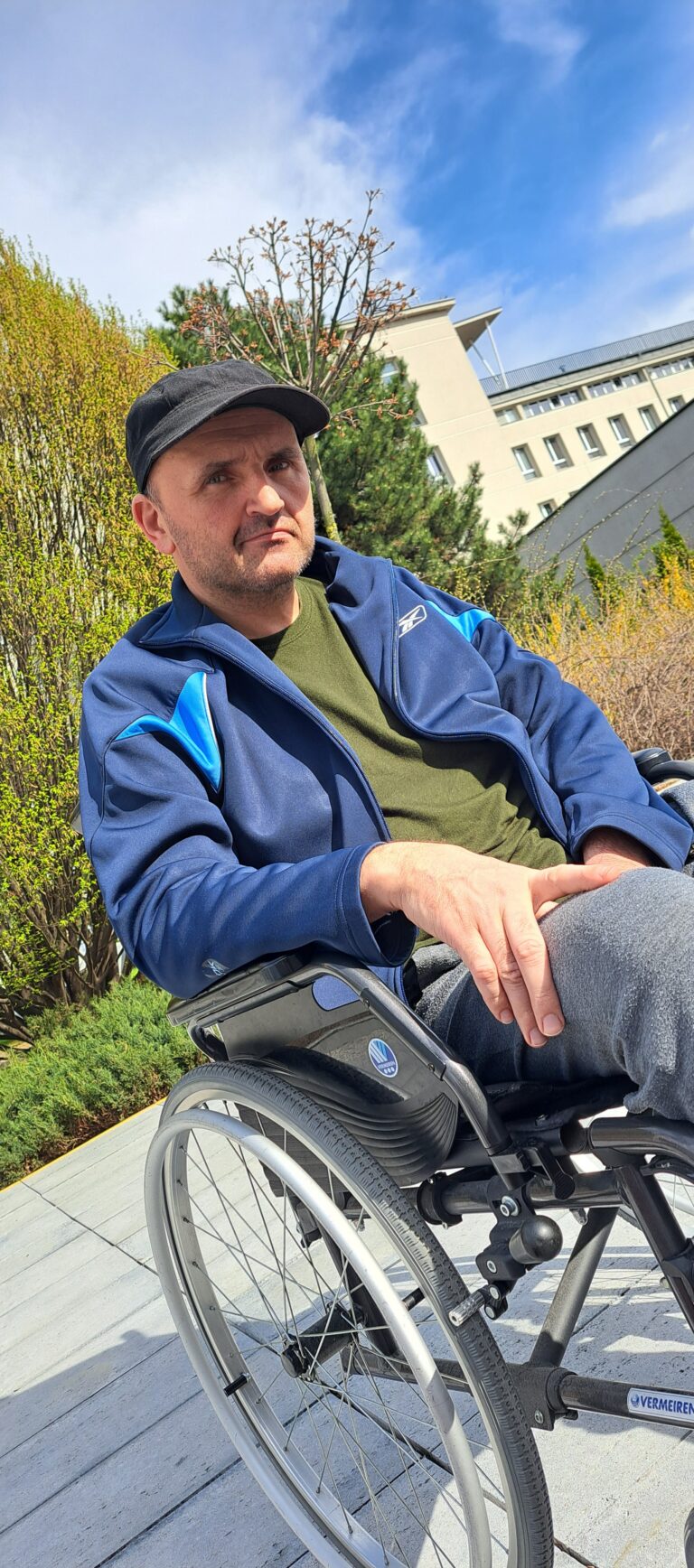 Adam Szawuła siedzi za wózku inwalidzkim i pozuje do zdjęcia. W tle szpital kliniczny.