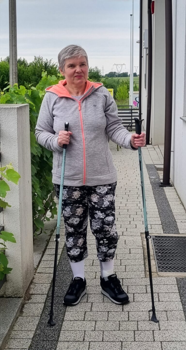 Teresa Zdunek wsparta o kijki do chodzenia pozuje przed budynkiem ośrodka neurorehabilitacji.