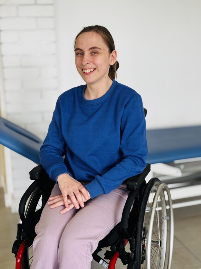 Anna Geneja uśmiechnięta pozuje do zdjęcia na wózku inwalidzkim w sali do rehabilitacji.