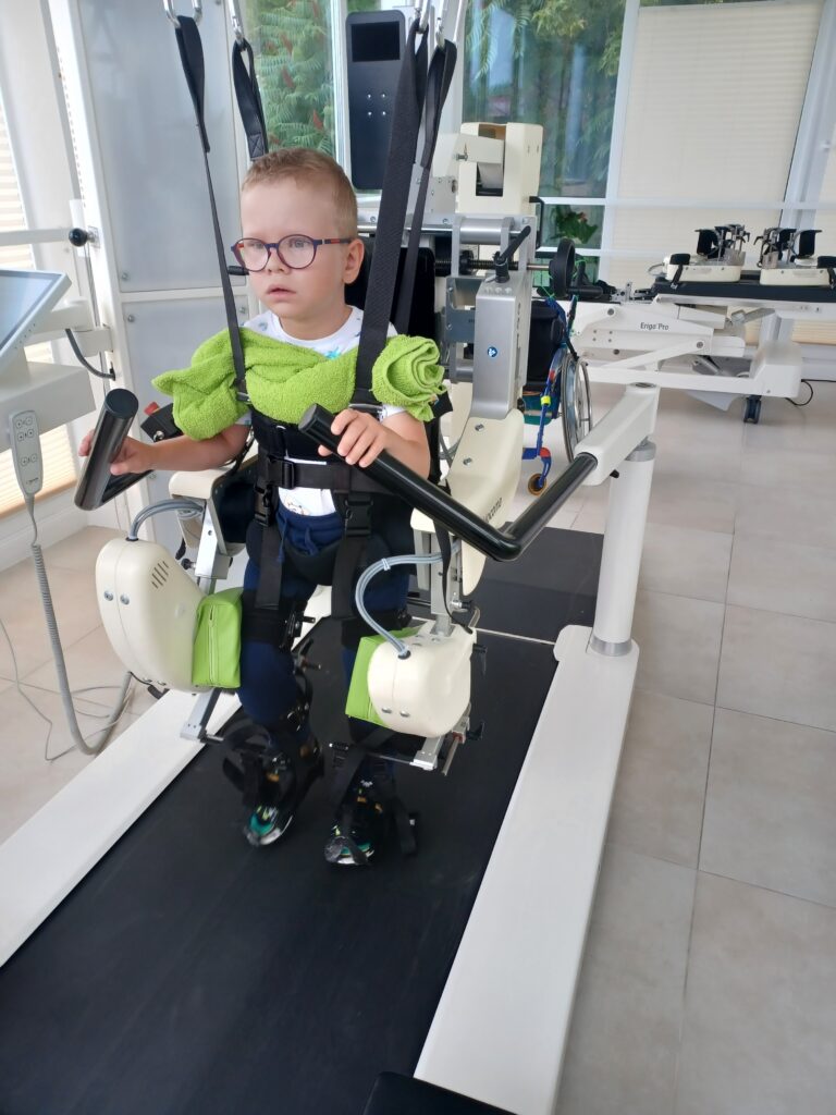Adrian Polajdowicz stoi zapięty w szelkach urządzenia do nauki chodu nad bieżnią.