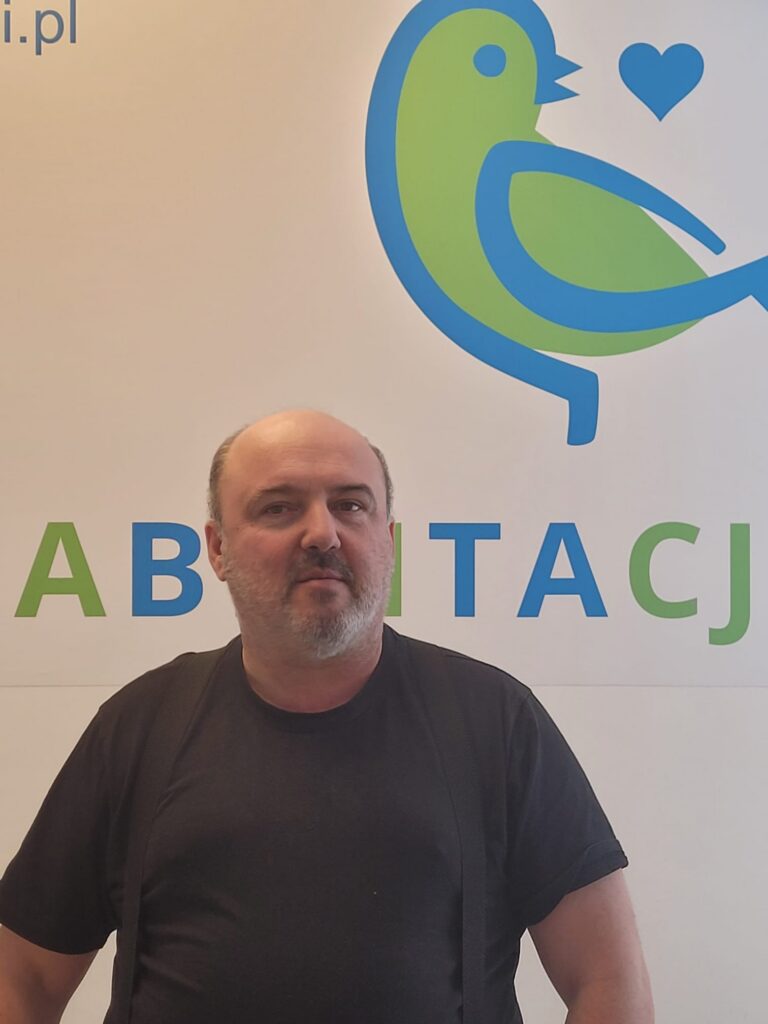 Andrzej Szlachta stoi przed ścianą z logo z ptakiem i napisem REHABILITACJA.