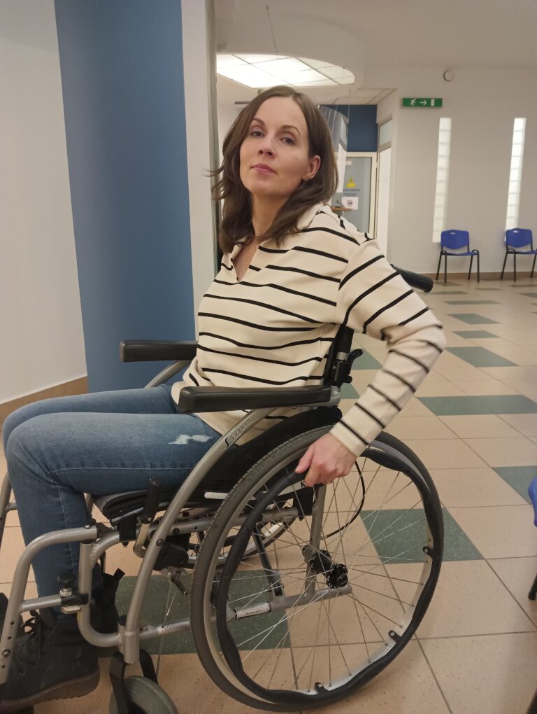 Ewa Szacon siedzi w wózku inwalidzkim w jasnym korytarzu, z dłonią na obręczy koła.
