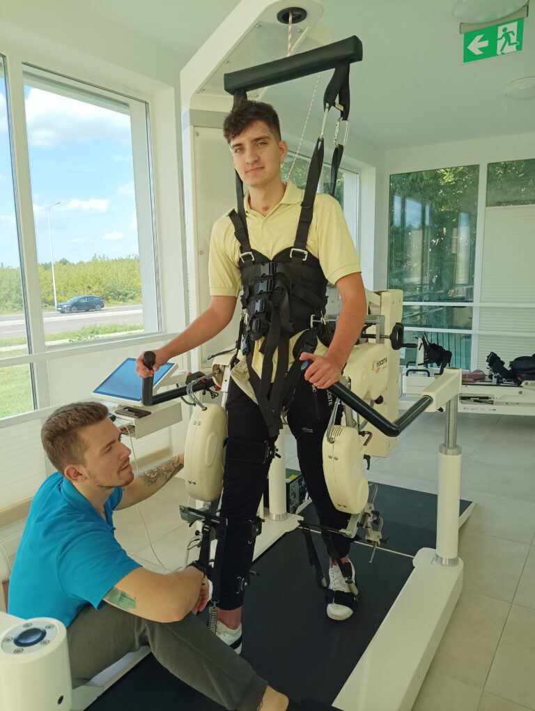 Franciszek Hołowiecki stoi zapięty w uprzęży nad bieżnią, na wysięgniku umieszczono tablet.