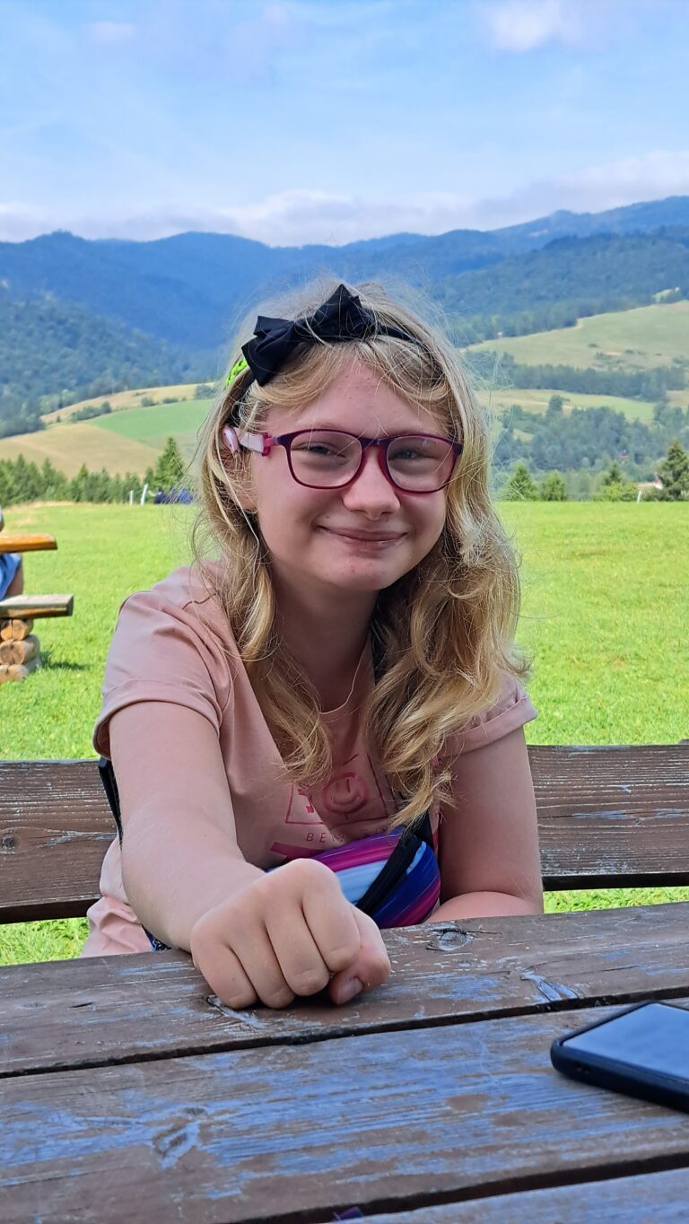 Iga Puzio opiera się o drewnianą barierkę, za nią rozciąga się łąka i wzgórza.