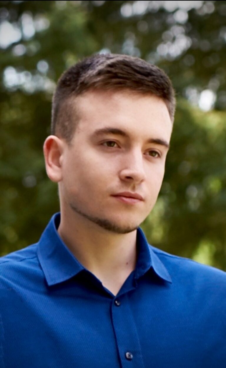 Kacper Lata patrzy w obiektyw, w tle rozmyta zieleń drzew.
