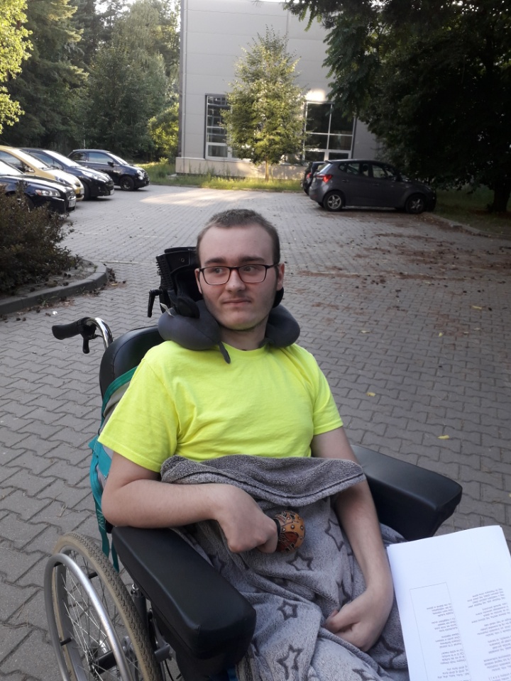 Kamil Fruchan siedzi w wózku na placu przed budynkiem i trzyma drewniany przedmiot.