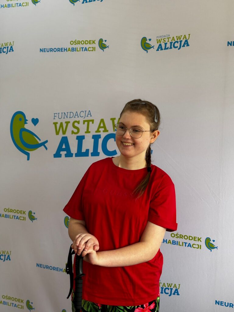Maja Kozłowska uśmiecha się, stojąc przy ściance z logo fundacji.