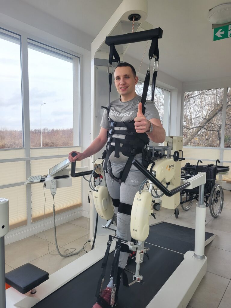 Marcin Kowalcze stoi w urządzeniu do nauki chodzenia i unosi kciuk w górę.