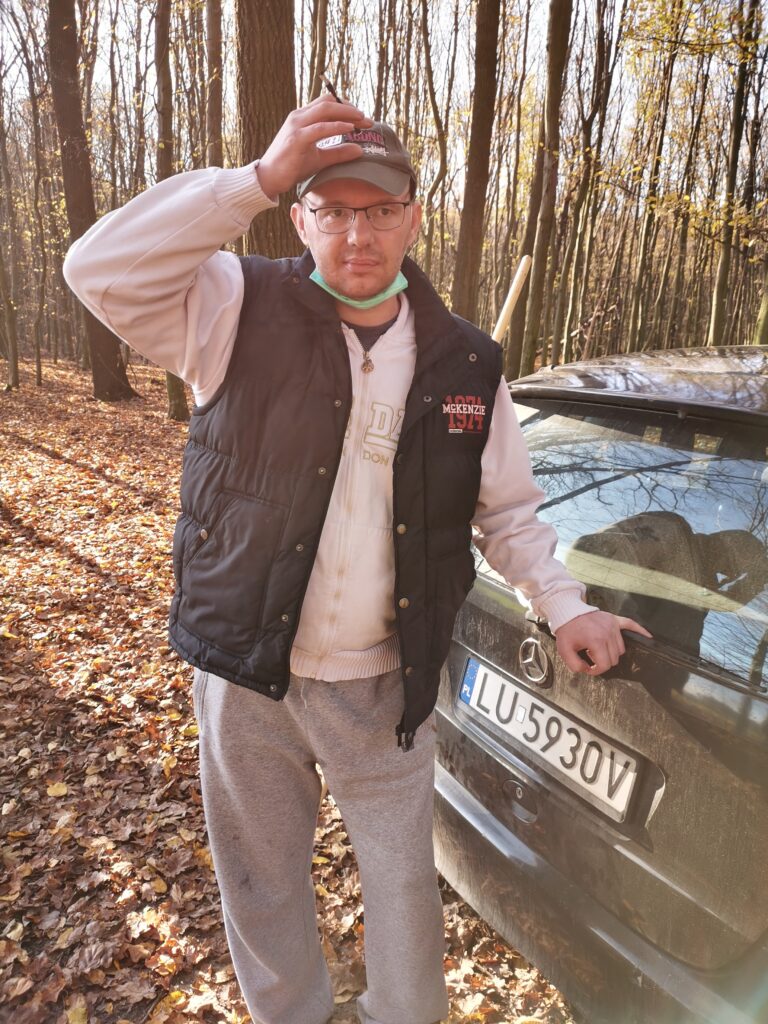 Mariusz Markiewicz stoi w jesiennym lesie i opiera rękę na samochodzie.