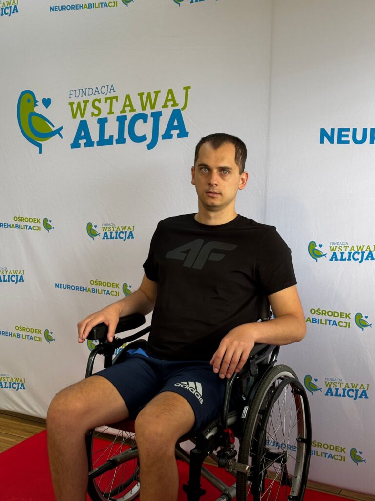 Michał Pacek siedzi w wózku przed ścianką z logo fundacji.