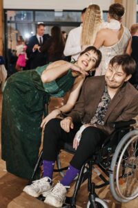 Tomek na wózku na sali weselnej, w brązowej marynarce i wzorzystej koszuli, żona pochyla się nad nim i śmieje.