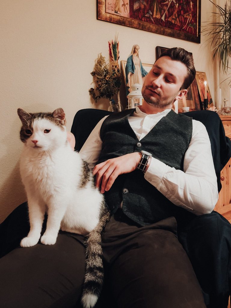Tomek w kamizelce i białej koszuli siedzi w fotelu, na kolanach ma biało-burego kota.